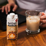 brüst cold brew latte protein coffee bottle next to a glass of iced coffee at a work desk