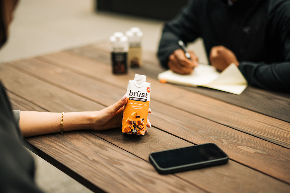 person holding a brüst salted caramel latte protein coffee bottle at a patio table